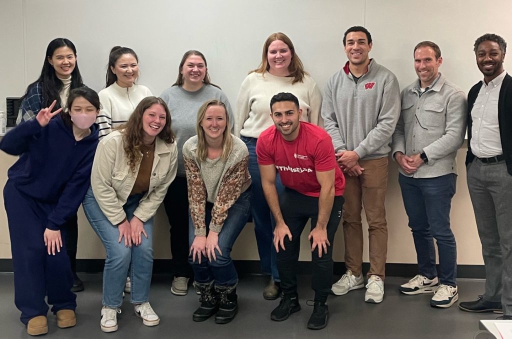 10 first year PhD students smile for a picture with Dr. Brian Burt in a classroom in the School of Education Building in front of a white board. The four students in the front row are doing a half squat. The students pictured were enrolled in Doctoral Inquiry in Fall 2024 taught by Dr. Brian Burt.