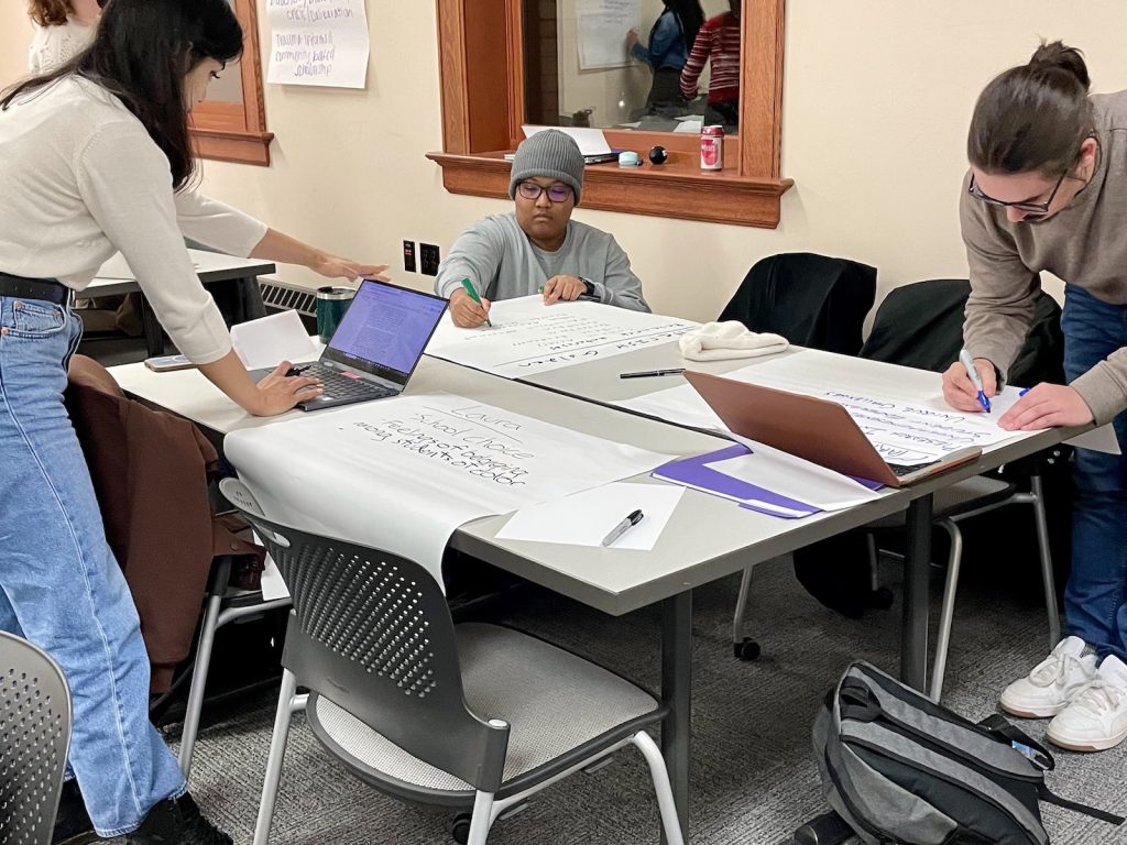 Three students in ELPA 824 work at a table on in-class work. One student is standing and reading from their laptop while the other two students write on large white post-it notes.