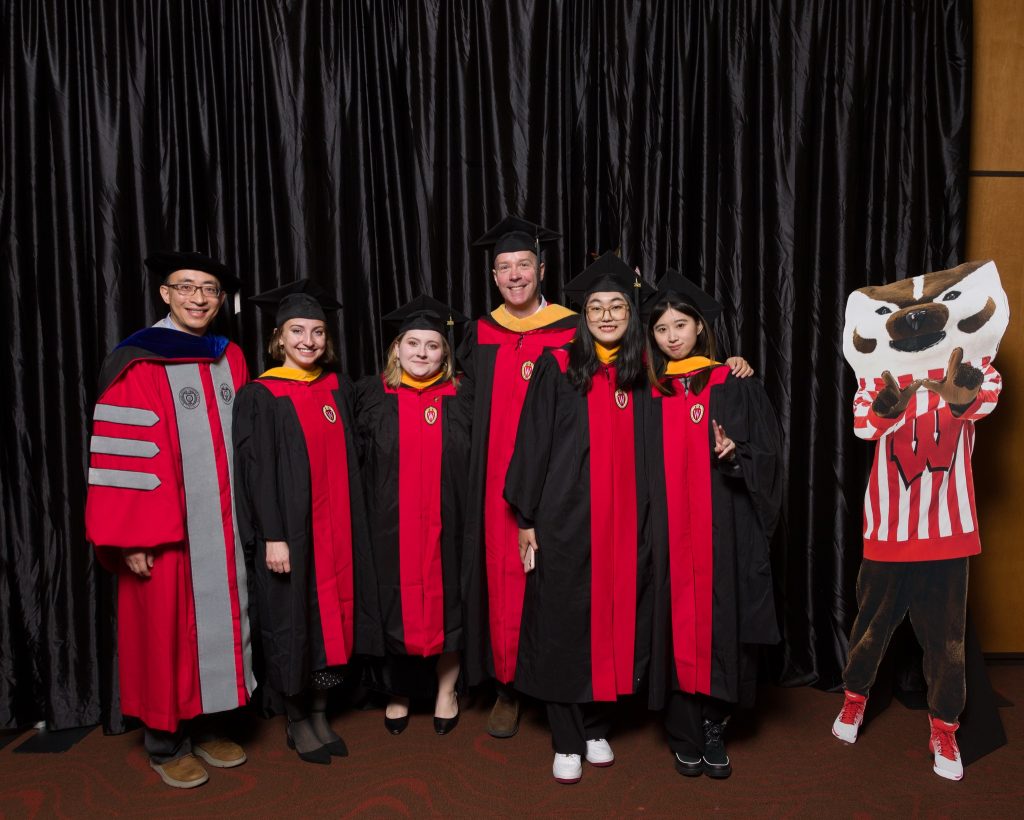 Five GHE students and Dr. Weijia Li smile for a group picture in their graduation robes in front of a black velvet curtain next to a cardboard cutout of Bucky Badger. The students were attending the School of Education's Winter 2024 Graduation Reception.