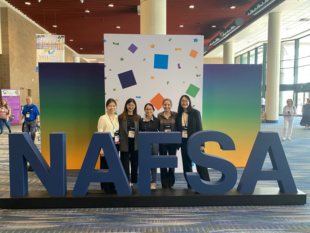Five GHE students attending the NAFSA conference stand and smile for a group photo in the conference photo area. Behind the students is a gradient backdrop and large letters spelling out "NAFSA" are in front of them.