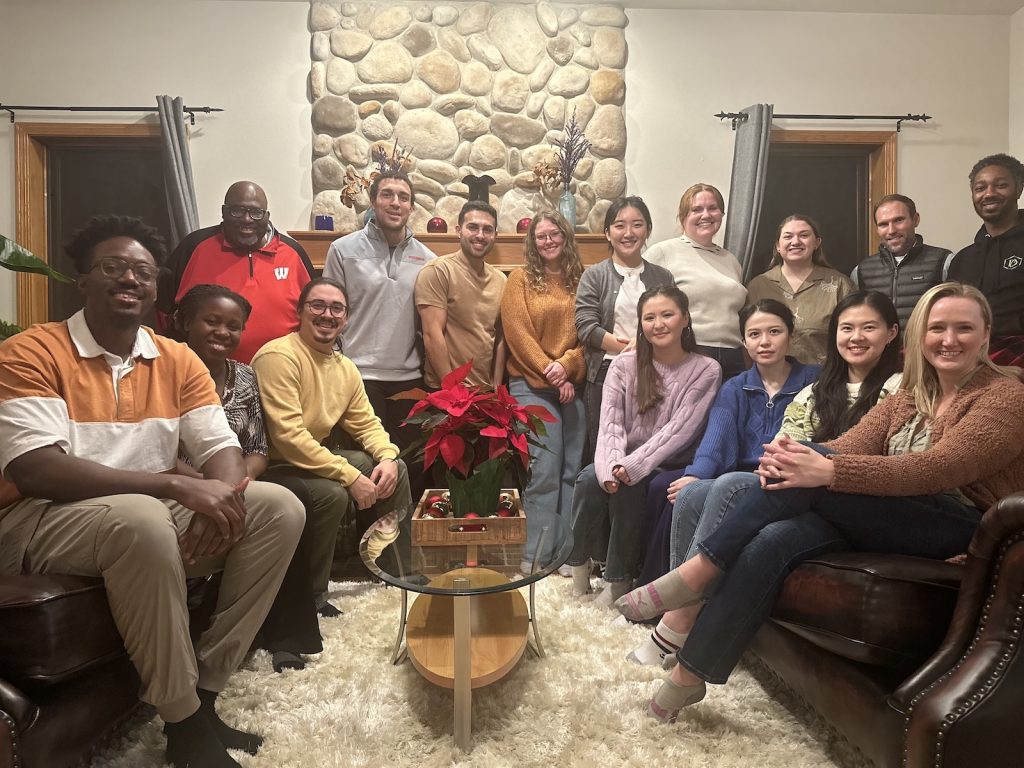 A group of 15 students and Dr. Brian Burt are sitting and standing in a semi-circle in a living room smiling for a group photo. The students include first year doctoral students that took Dr. Burt's Doctoral Inquiry course in Fall 2024 and Dr. Burt's advisees.