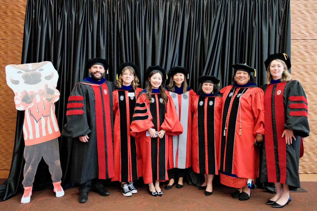 From left to right: Joseph Romero-Reyes, Ayse Okur, Peiwen Zheng, Dr. Xueli Wang, Valerie Crespin-Trujillo, Nicole Soulier, and Maria Widmer smile for a group photo at Spring Graduation 2024. Everyone is wearing their graduation robes and are standing next to a cardboard cutout of Bucky Badger. The photo is set in front of a black velvet curtain at the School of Education Graduation Reception.