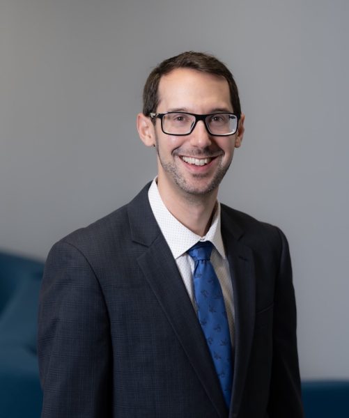 A headshot of Dr. Brett Ranon Nachman, who is smiling in a gray room with a blue chair in the lower left corner.