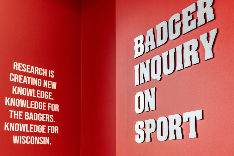 A red wall in the BIOS lab with the lab name spelled out in big letters, "Badger Inquiry On Sport." To the left of the lab name, the following phrase is painted on the wall, "Research is creating new knowledge. Knowledge for the Badgers. Knowledge for Wisconsin."