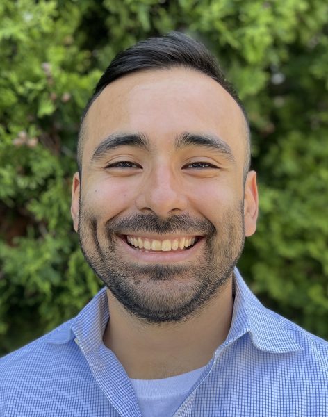 A headshot of Dr. Daniel (Danny) Corral, who is smiling in front of a green foliage wall.