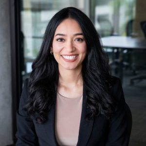 A headshot of Dr. Nicole Contreras-García, who is sitting with her hands crossed and smiling in front of a School of Education Building glass wall.