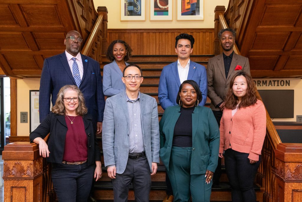 The 2024-25 Higher Education faculty posing for a picture on the main staircase inside the School of Education Building. They are standing in two rows of four and smiling at the camera