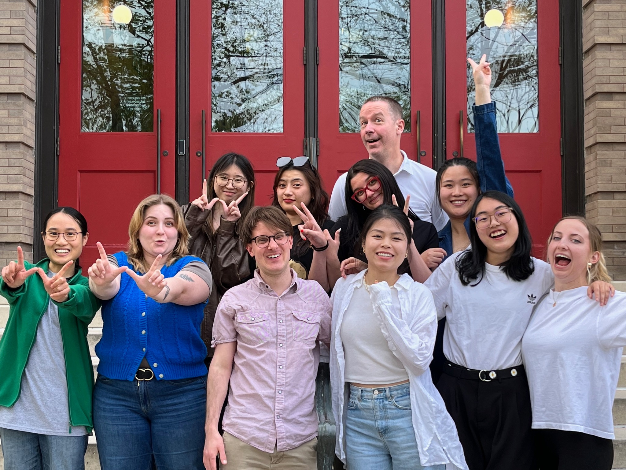 11 students from the 2023 GHE cohort pose for a fun picture on the steps of the School of Education Building in front of the red doors. Some of the students are smiling, some are making silly faces, and some are laughing. A couple of students are making the Wisconsin 