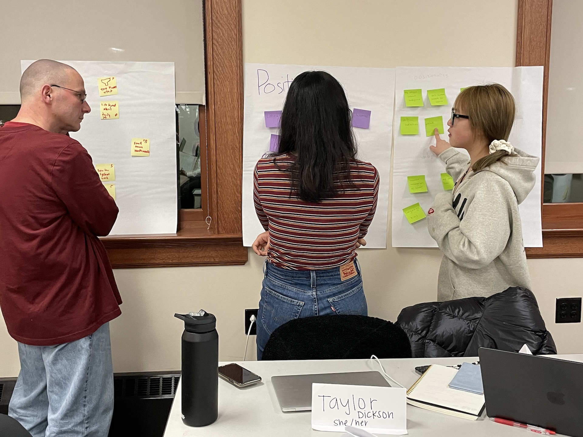 Three students in ELPA 824 work together on an in-class group assignment. The students are facing a window that is covered in three large white post-it notes that have smaller post-it notes on them.