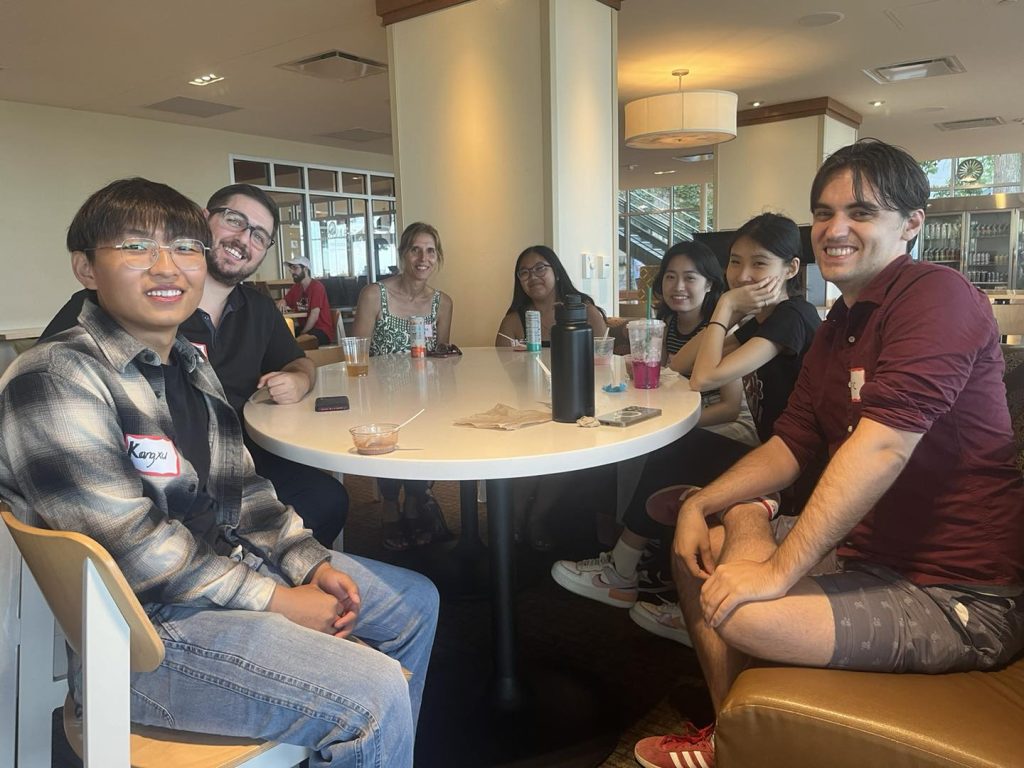 Seven students from the GHE 2024 cohort are sitting in a semi-circle around a table inside the Memorial Union and smiling for a group photo. A variety of drinks and empty Babcock Ice Cream dishes sit on the table.