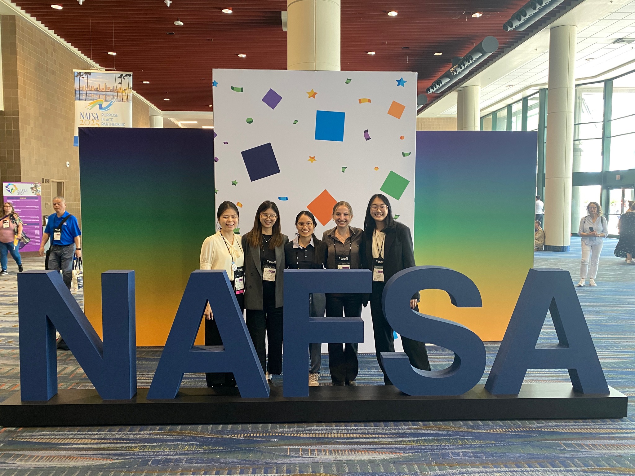 Five GHE students attending the NAFSA conference stand and smile for a group photo in the conference photo area. Behind the students is a gradient backdrop and large letters spelling out 