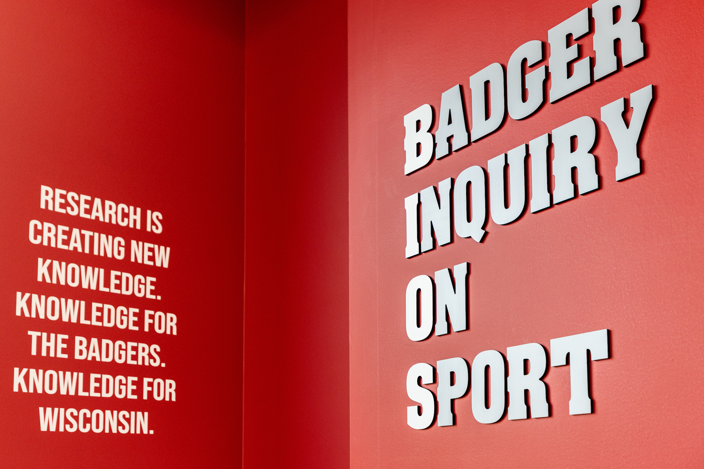A red wall in the BIOS lab with the lab name spelled out in big letters, "Badger Inquiry On Sport." To the left of the lab name, the following phrase is painted on the wall, "Research is creating new knowledge. Knowledge for the Badgers. Knowledge for Wisconsin."