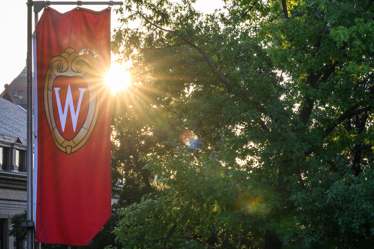 A red banner featuring the UW–Madison crest is featured in the foreground on the left side of the image, with the sunset peaking through a green tree in the background.