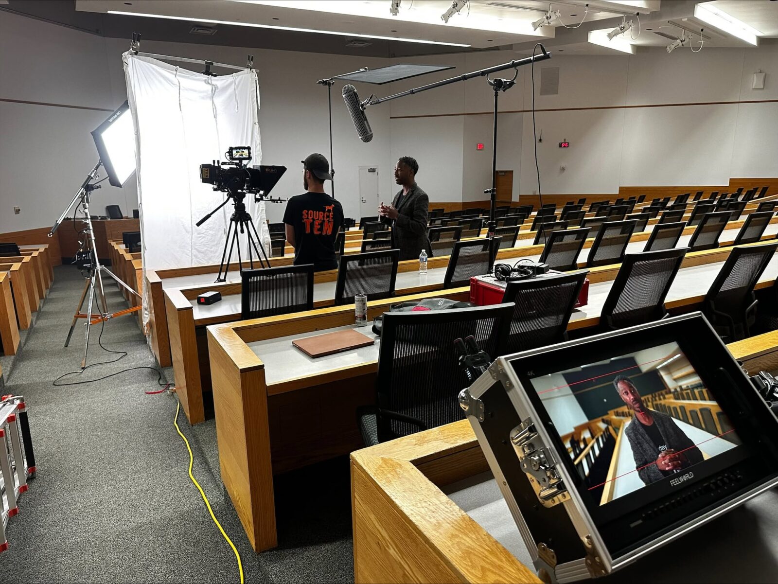 Dr. Brian A. Burt with a camera person in a lecture hall filming for a video project. There is a white background screen and a boom microphone near Dr. Burt and the camera person.