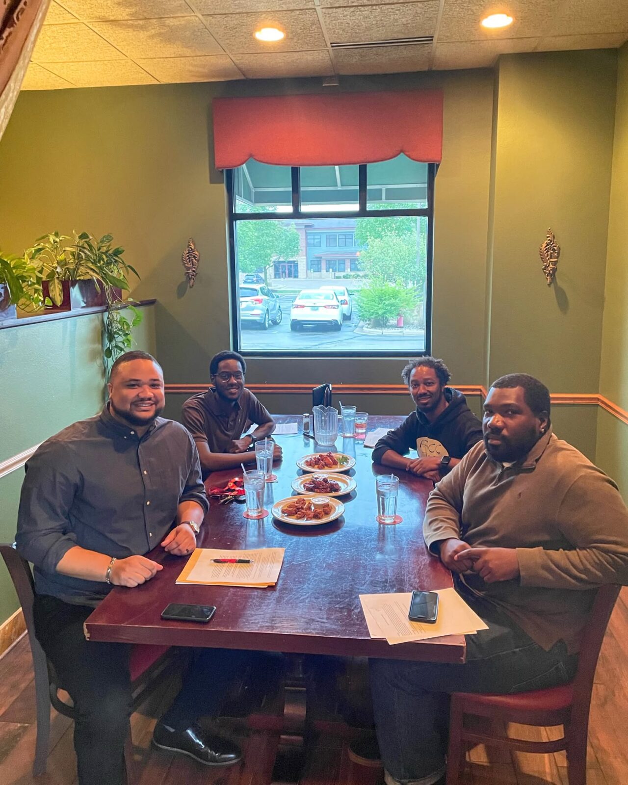 Dr. Brian A. Burt sits around a table with advisees and all smile at the camera. From left to right: (now) Dr. Joshua Wallace, Devin Henry, Dr. Burt, and (now) Dr. Blayne Stone, Jr.