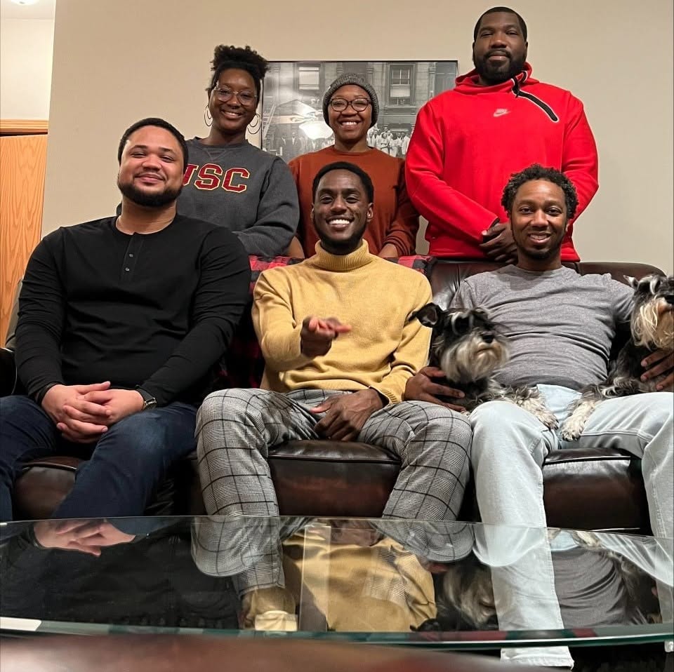 Dr. Brian A. Burt sits on a couch for a group photo with members of the Wei Lab. Back row, standing left to right: Dr. Dajanae Palmer, (now Dr.) Tiaira Porter-Beall), (now Dr.) Blayne Stone, Jr.); front row, sitting on the couch: (now Dr.) Joshua Wallace, Devin Henry, Dr. Burt (with a dog on each leg).