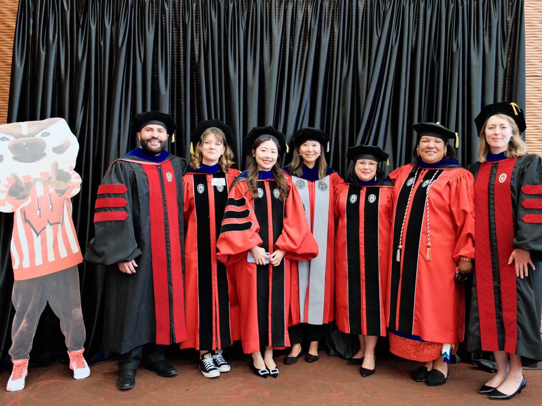 From left to right: Joseph Romero-Reyes, Ayse Okur, Peiwen Zheng, Dr. Xueli Wang, Valerie Crespin-Trujillo, Nicole Soulier, and Maria Widmer smile for a group photo at Spring Graduation 2024. Everyone is wearing their graduation robes and are standing next to a cardboard cutout of Bucky Badger. The photo is set in front of a black velvet curtain at the School of Education Graduation Reception.