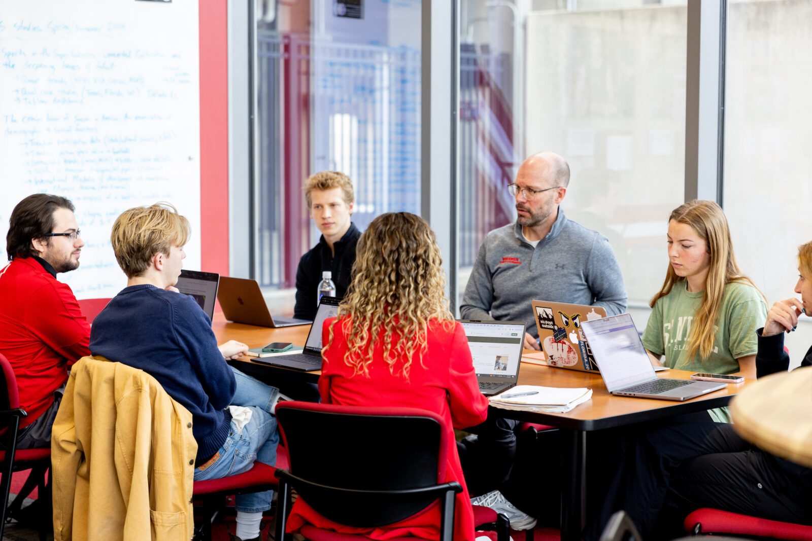 Dr. Pete Miller, Maria Dehnert, and Dr. Sara Jimenez Soffa sit around a table with students in the BIOS Research Lab working on their laptops and discussing their work.