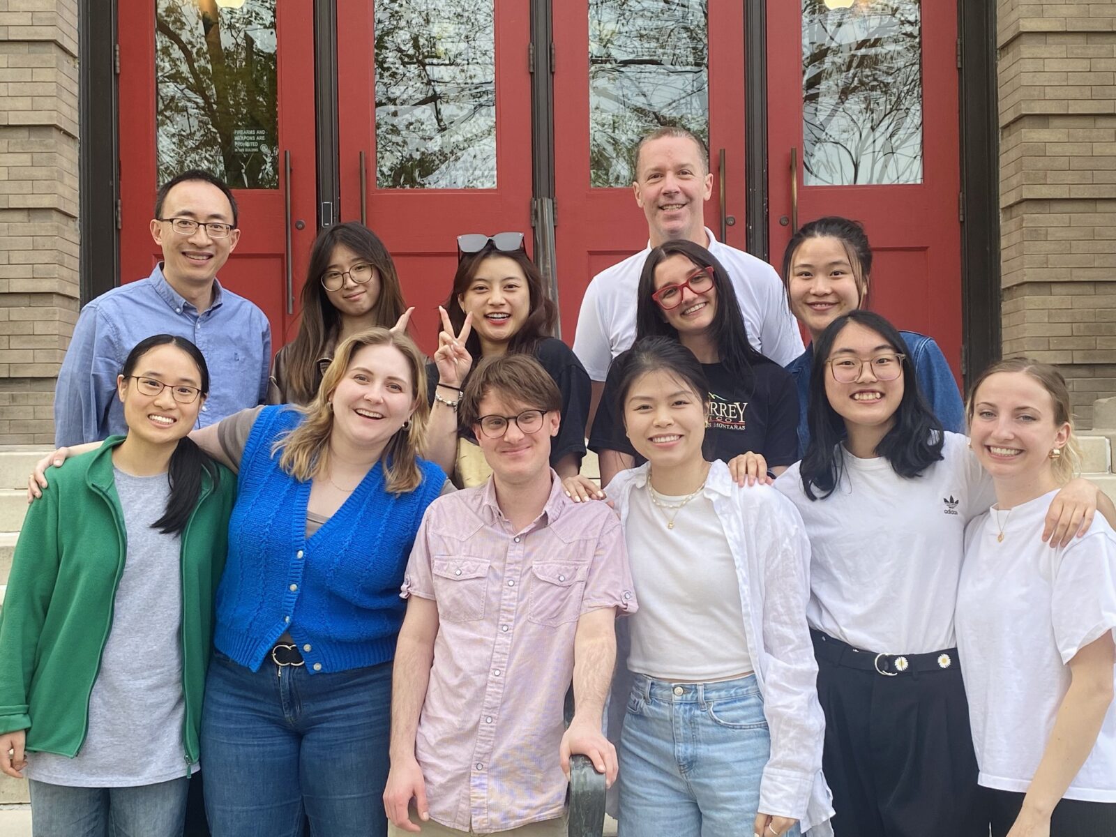 11 GHE master's degree students smile in a group photo with Dr. Weijia Li on the stairs of the Education Building in front of the Red Doors.