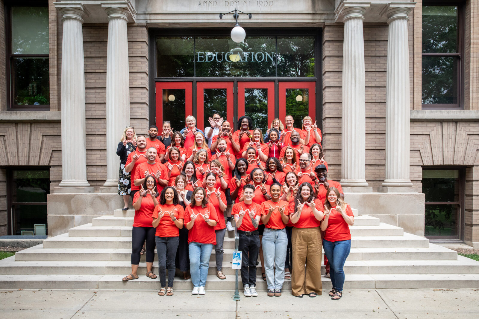 The MS K-12 Leadership and DLPP Cohorts smile and make the UW–Madison "W" with their hands, along with ELPA and School of Education faculty. The group is standing on the stairs of the Education Building in front of the red doors.