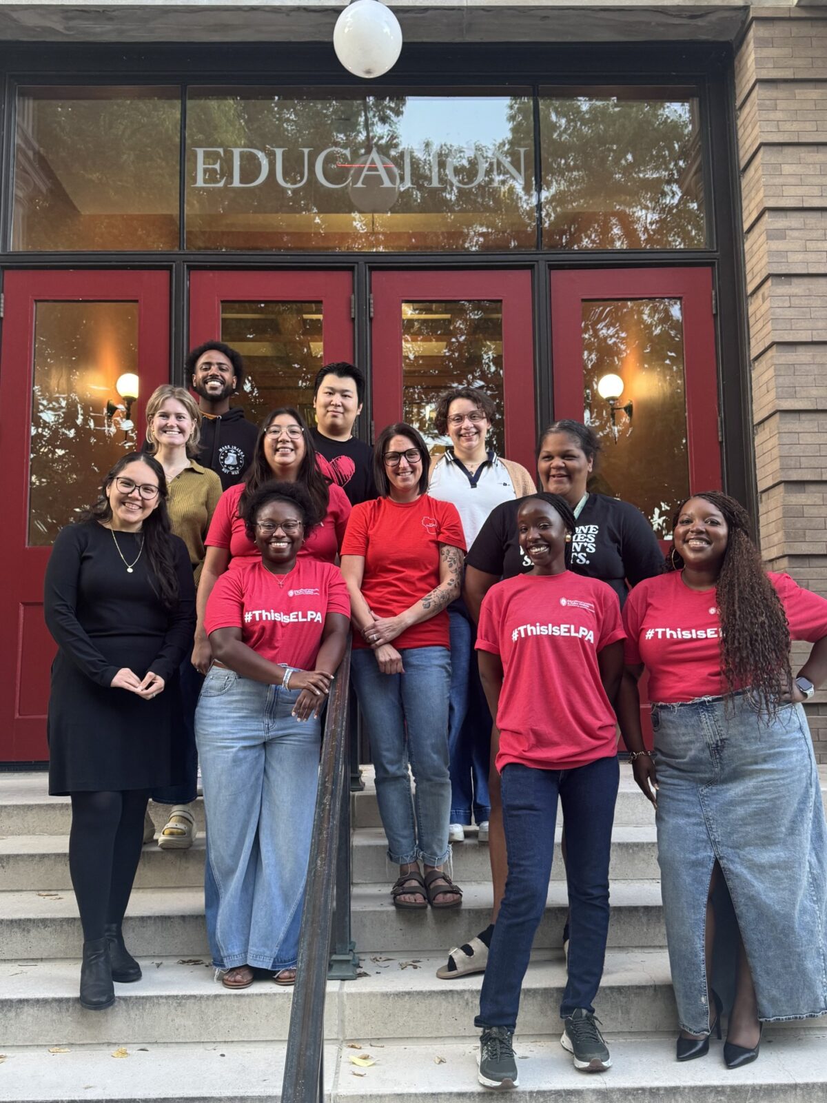 A group of 10 first-year ELPA PhD students are standing together and smiling with Dr. Aireale Rodgers on the stairs of the Education Building in front of the iconic red doors. Some of the students and Dr. Rodgers are wearing #ThisIsELPA red t-shirts.