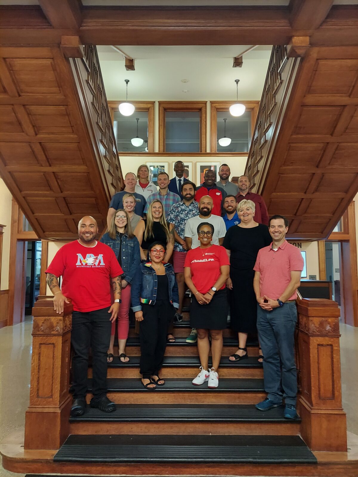 A group of 18 adults standing scattered on the main staircase in the School of Education building and smiling at the camera. The group features 16 students in the WIE PhD Cohort, program director Dr. Barb Sramek, and former ELPA Department Chair Dr. AJ Welton.