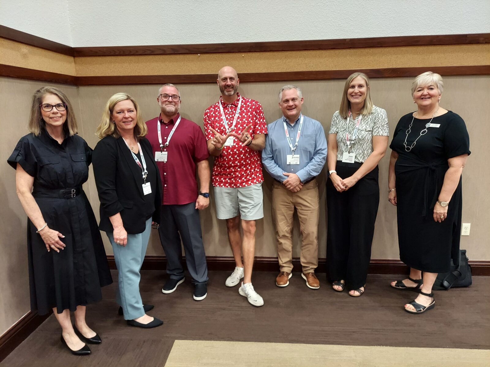 Six ELPA alumni and Dr. Barb Sramek (also an alum) stand in a semi-circle in front of a wall and smile for a picture. Dr. Neil O'Connell is in the middle of the group, holding up the Wisconsin “W” with his fingers.