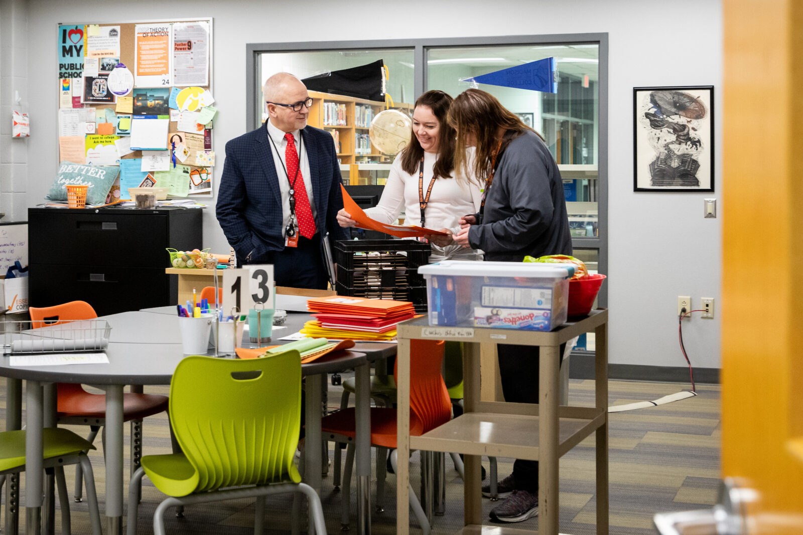 An ELPA student at Oregon High School looks at an orange folder with her principal and another administrative staff member in a colorful office space by a table.